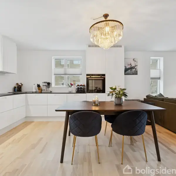 Køkken med hvidt interiør i aktion, der støder op til en spiseplads med sort bord og to stole. En lysekrone hænger over bordet, sofaen i baggrunden under vinduer.