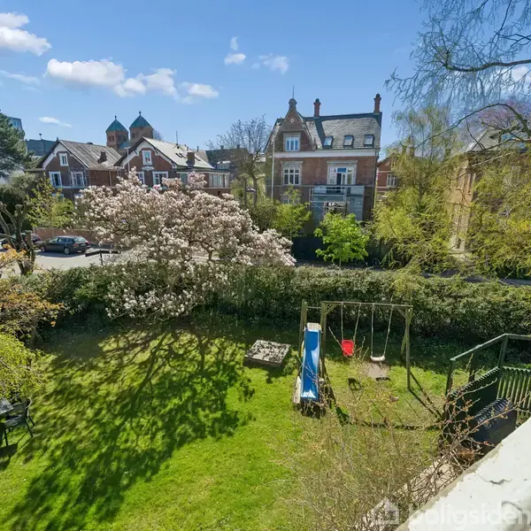 En blå rutsjebane står på en grøn græsplæne, omgivet af blomstrende træer og buske. En træterrasse med potteplanter fører ud i haven, og et hus er i baggrunden.