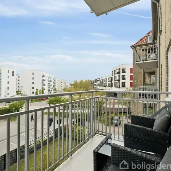Altan med sort fletmøblement, hvorfra man kan overse en moderne boligblokbygning og vej. Solrigt vejr og blå himmel danner baggrund til denne bypanoramaudsigt.