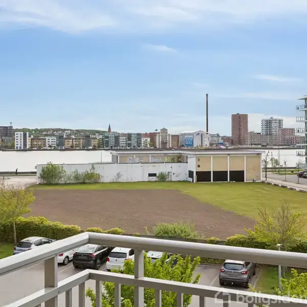 Balkon med udsigt over parkeringsplads og græsareal. I baggrunden er der moderne lejlighedsbygninger og industribygninger. Klar himmel med lette skyer.