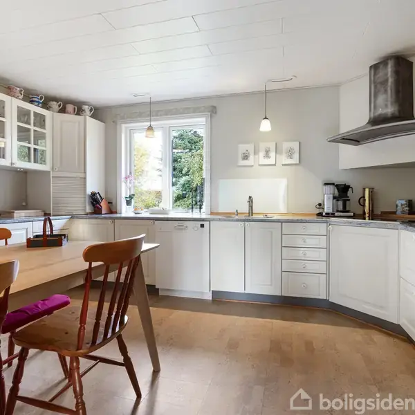 Køkken med hvide skabe og indbyggede apparater; et træbord med stole står i midten. To lamper hænger i loftet, og et vindue giver udsigt til en have.