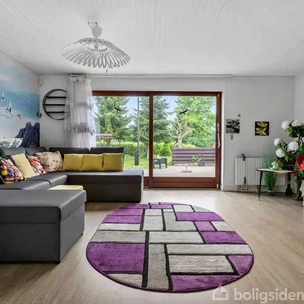 Sofa i gråt læder står foran en stor vindue, der viser en grøn have. Et lilla tæppe ligger på gulvet. Der er et farverigt vægmaleri og en sort vægmonteret TV.