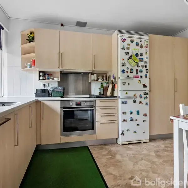 Et køkken med lyse træskabe og en indbygget ovn. Køleskabet, dekoreret med magneter, står ved siden af et bord og stole. Et vindue lader lys komme ind.