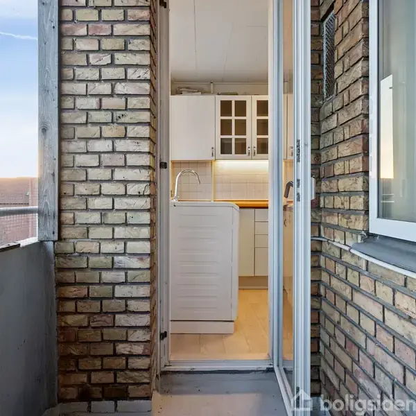 Balkon med murstensvægge fører til et åbent køkken med hvide skabe. Et vindue ses til højre, og balkonen har udsigt over et byområde med himlen i baggrunden.