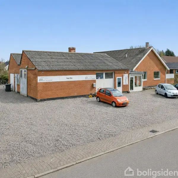 Rød murstensbygning med fladt tag står på en grusparkeringsplads. Tre biler er parkeret foran. Skilte på bygningen viser "Hundehårene" og "Salon pelspleje".