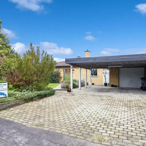 Garage med fladt tag står på brostensbelagt indkørsel i villakvarter. Et lille skilt viser teksten "Til salg". Huset med gule mursten er omgivet af grøn beplantning.