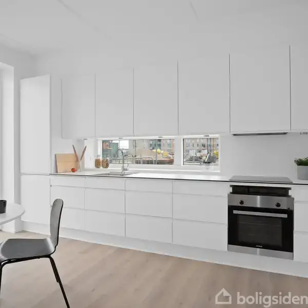 Hvidt køkken med indbyggede skabe og en ovn. Bordplade med vask under et bredt vindue. Runde spisebord med sorte stole. Lys træparket og glasdør til terrasse.