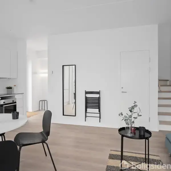 En moderne stue med et rundt spisebord med stole, pyntet med røde dekorative grene. En hvid væg har et spejl og en sort stol. Trappe til højre.