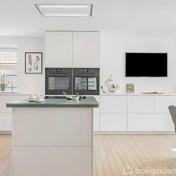 Spisebord med sorte lænestole står i et lyst køkken. Store vinduer giver naturligt lys, og et moderne, hvidt indretningsdesign dominerer rummet. En sort vægmonteret skærm overfor bordet.