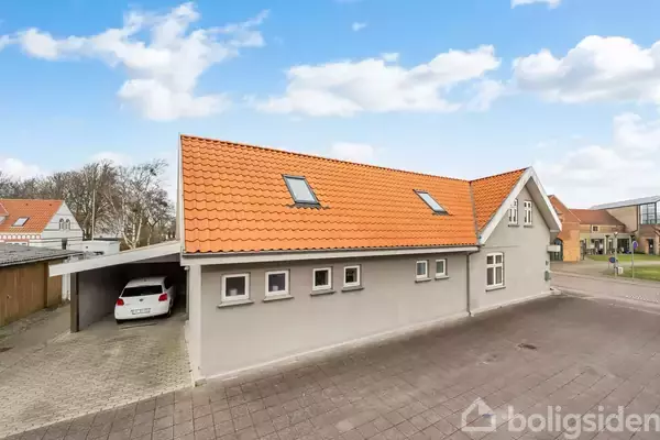 Huset har et orangerødt tegltag og grå vægge, parkeret bil i carporten. Det står i et boligområde med enkeltstående træer og skyet blå himmel.