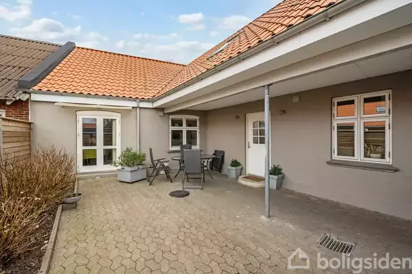 En gårdhave med fliser og havemøbler står klar til brug. Omkranset af et hus med rødt tegltag og hvidrammede vinduer giver det en hyggelig atmosfære.