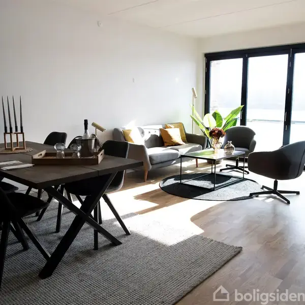Spisebord med sorte stole står på et tæppe i en stue med sofa, sofabord og lænestole. Sollys kommer ind gennem store vinduer ved siden af et hyldesystem med planter.