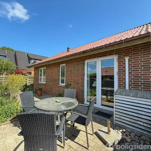 Et rundt havebord med sorte fletstole står på en terrasse foran et murstenshus med rødt tegltag. En have omringer området med grønne planter og hegn.