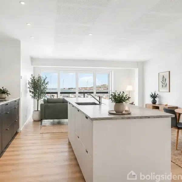 Køkken med hvid køkkenø og indbyggede apparater, parallelt med træskabe på venstre side. Stue med grå sofa og store vinduer, ved siden af spisebord med lamper.