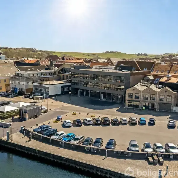 Parkeringsplads med biler arrangeret i rækker ved en havn. Baggrund består af bygninger med teglstenstage og en kirke i landsbyen under klar himmel.