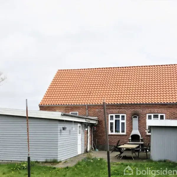 Et rødt murstenshus med orange tag og to vinduer, omgivet af en grøn græsplæne. En udendørs pejs og havemøbler står i gården ved siden af et gråt skur.