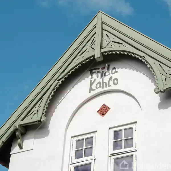 Et hvidt hus med grønne gavle har teksten "Frida Kahlo" over vinduerne. Det er en solrig dag med blå himmel i baggrunden.