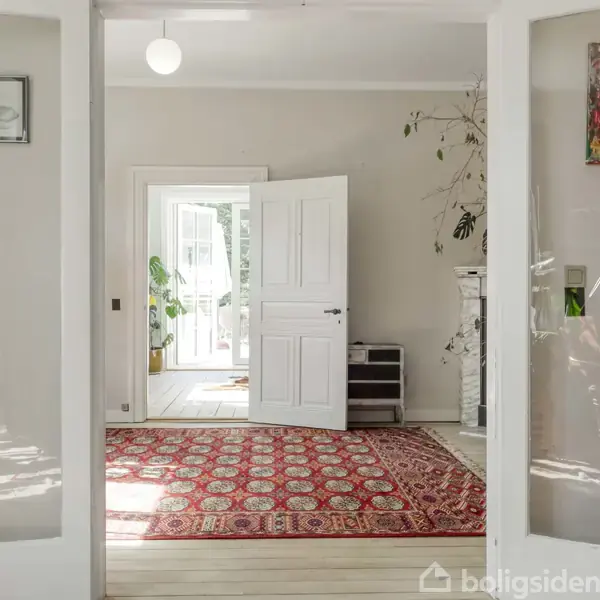 Gennemsigtige dobbeltdøre fører ind til en stue med et orientalsk tæppe på trægulv. Et åbent vindue i baggrunden tillader dagslys. Væggen har en farverig kunstplakat.