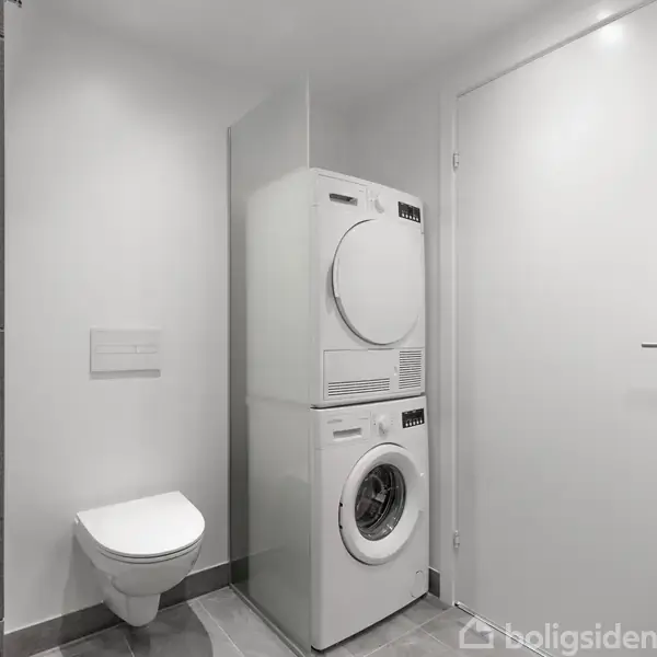 Vaskesøjle med vaskemaskine og tørretumbler er placeret ved siden af hvid dør i badeværelse med grå fliser; væghængt toilet findes til venstre for søjlen.