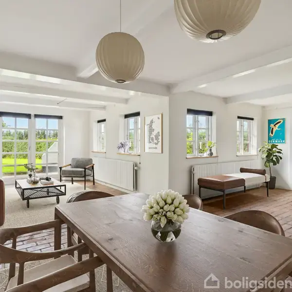 Et rummeligt stueområde med et spisebord i træ forrest omgivet af stole. Lædermøbler og planter ses i baggrunden, med store vinduer, der slipper naturligt lys ind. En bogreol står ved siden af en plante.