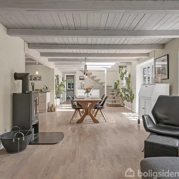 Stue med brændeovn til venstre, spisebord i midten, og moderne stol til højre. Rummet er lyst med træbjælker og planter, mens en trappe leder op i baggrunden.