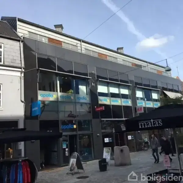 En moderne bygning med glasfacade indeholder butikker; vinduer viser navne som "apollo" og "matas". Folk går forbi på en brostensbelagt gade med udendørs parasoller og butiksskilte.