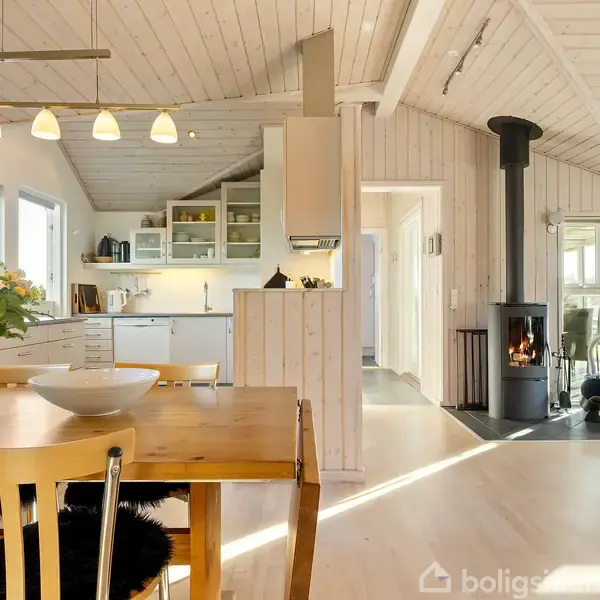Træspisebord med blomster og skål står i åben stue. Pejs brænder, køkken i baggrunden. Lyse trævægge og loft skaber en hyggelig atmosfære.