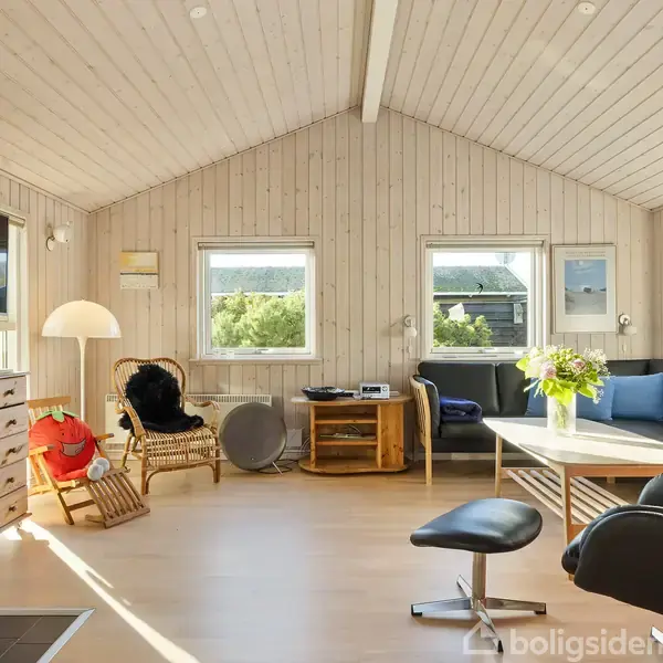 Stue med lyse træpaneler indeholder en sofa med puder, et sofabord med blomster, en lænestol og en fladskærm på væggen. Sollys strømmer ind gennem vinduerne.