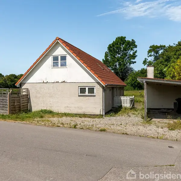 Et lille hus med rødbrunt tegltag står ved en vej. Huset har et hegn og en carport til højre. Grønt landskab omgiver bygningen, og det er en solrig dag.