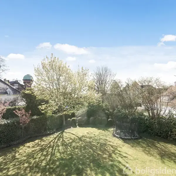 Græsplæne badet i sollys, omkranset af frodige buske og træer. Nabohuse kigger frem over hækken, mens en blå, skyfri himmel spreder sig i baggrunden.