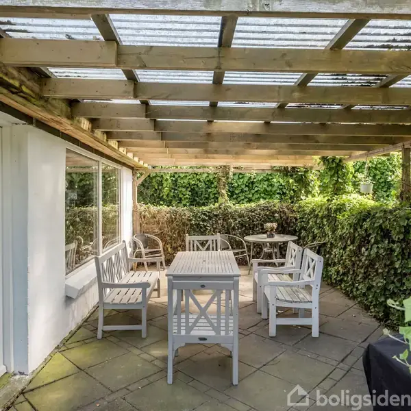 Terrasse med hvidmalede træmøbler under et pergolatak af glas. Bord og stole står på flisebelagt gulv. Omkring er der tæt, grøn beplantning, hvilket skaber en intim, indbydende atmosfære.