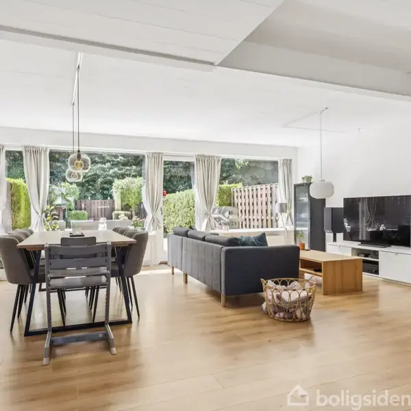 En moderne stue med trægulv indeholder spisebord med stole og en sofa ved siden af et TV og mediemøbler. Store vinduer med gardiner åbner til en have.