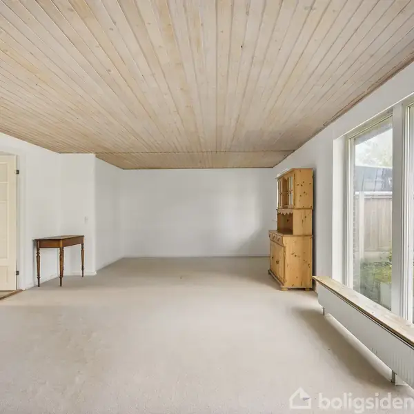 Et rummeligt, lyst værelse med trægulv og træloft. Et lille bord står ved en åben dør til venstre, mens en træskænk står til højre. Store vinduer vender ud mod en have.