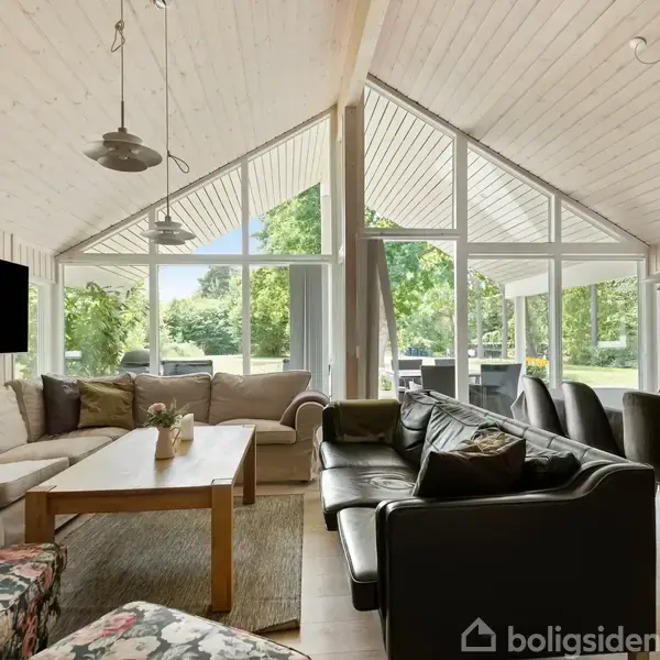 En stue med sofaer og et sofabord er centralt placeret. Store vinduer giver udsigt til en frodig have. Et spisebord står til højre, og loftlamper hænger ned.