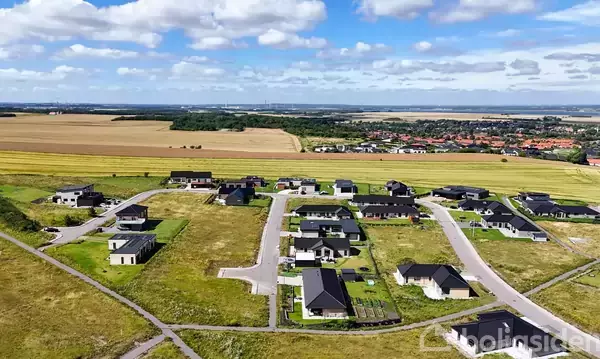 Boligområde med moderne huse ligger på grønne, åbne marker. Små veje forbinder husene, mens marker og en by ses i baggrunden under en blå himmel med skyer.