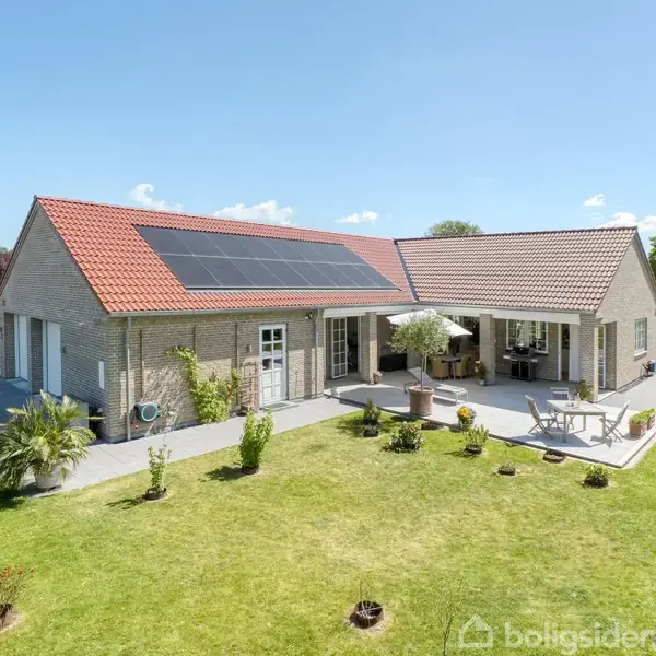 Et moderne enfamiliehus med solpaneler på taget, ligger på en velplejet græsplæne. Huset har en terrasse med havemøbler, omgivet af en række buske og hegn.
