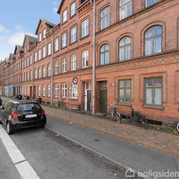 En murstensbygning med buede vinduer står langs en byvej. Cykler er parkeret ved siden af fortovet. Biler kører på vejen. En vejskilt viser "Indkørsel forbudt".