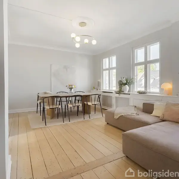 Stue med sofa til højre og spisebord med stole til venstre. Rummet har trægulv, store vinduer og en moderne lampe i loftet. Skandinavisk indretning med neutrale farver.