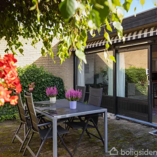 En terrasse med et bord og fire stole står i forgrunden. Blomster står på bordet. I baggrunden ses en glasdør, der fører til en stue med spisebord og stole.