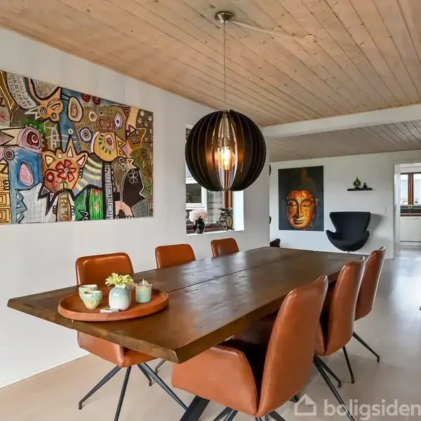 Langt træbord omgivet af læderstole står i spisestuen, dekoreret med moderne kunst på væggene og en stor pendellampe over bordet. En fjern stue kan ses i baggrunden.