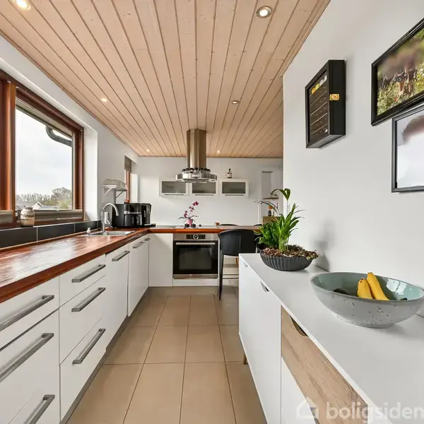 Køkken med hvide skabe og træbordplade, indeholder kogeplade, ovn og emhætte. Rum med store vinduer, flisegulv og billeder på væggen; skål med bananer på bordet.