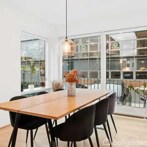 Et spisebord med seks sorte stole er placeret nær vinduer, hvor lys trænger ind. Et rundt spejl hænger på væggen, og en lampe hænger over bordet.