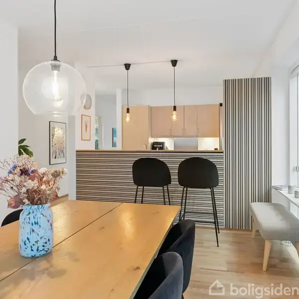 Et spisebord med en blå vase og blomster står i et lyst køkken-alrum. To sorte barstole står ved køkkenøen, omgivet af lyse moderne lamper og træmøbler.