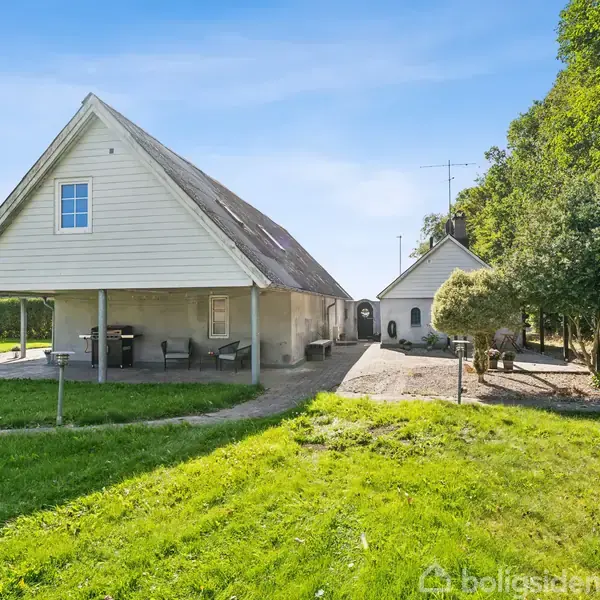 En lysegrå bygning med stejl taghældning står stille i en frodig have, der er omgivet af grønne træer, med en flisebelagt terrasse foran.