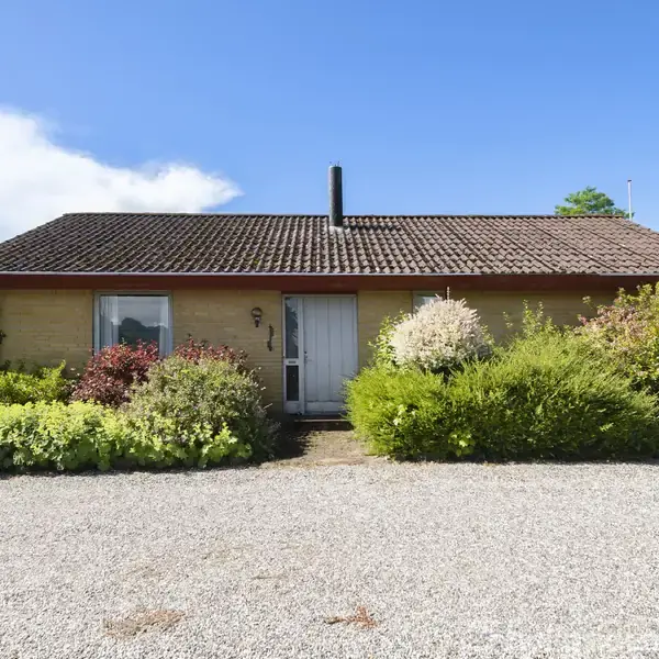 Et enfamilieshus med rødt tegltag står midt i en have med grønne buske og farverige blomster. Foran huset er der en grusbelagt indkørsel.