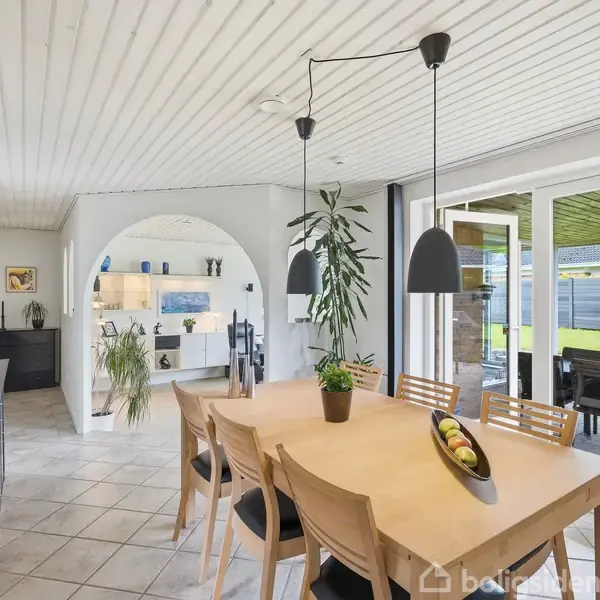 Et spisebord med træstole står under to sorte hængelamper. Foran er en stor glasdør med udsigt til en terrasse. I baggrunden ses en bueformet døråbning til en opholdsstue.