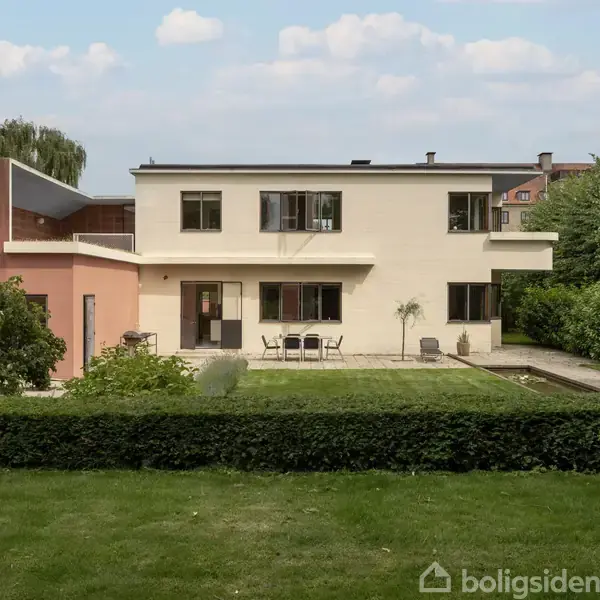 En moderne, hvid tofamiliersvilla står i en grøn have med en terrasse foran. Græsplænen er omgivet af hække og buske, mens himlen er blå med få skyer.