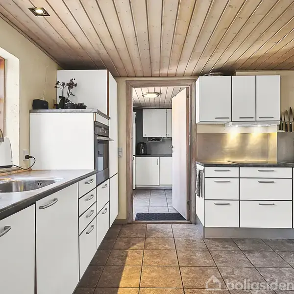 Køkken med hvide skabe og skuffer, grå bordplader, moderne vask, og ovn. Rummet har træloft og flisegulv. Et vindue giver lys, og der er redskaber hængende på væggen.