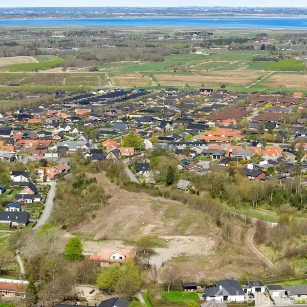Byområde strækker sig med mange huse med røde tage i forgrunden. Omkringliggende grønne marker og skove med en stor fjord i horisonten.
