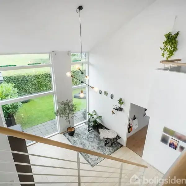 Trappe fører ned til en stue med store vinduer, der giver udsigt til en græsplæne. En stor plante, sofa og tæppe findes i hjørnet. Væggene er hvide med dekorative elementer.